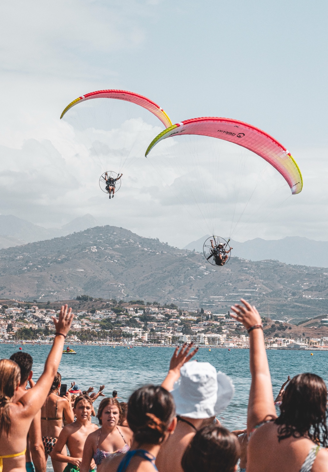 Dos pilotos de paramotor con velas de colores vuelan junto a la costa, ante el público en la playa